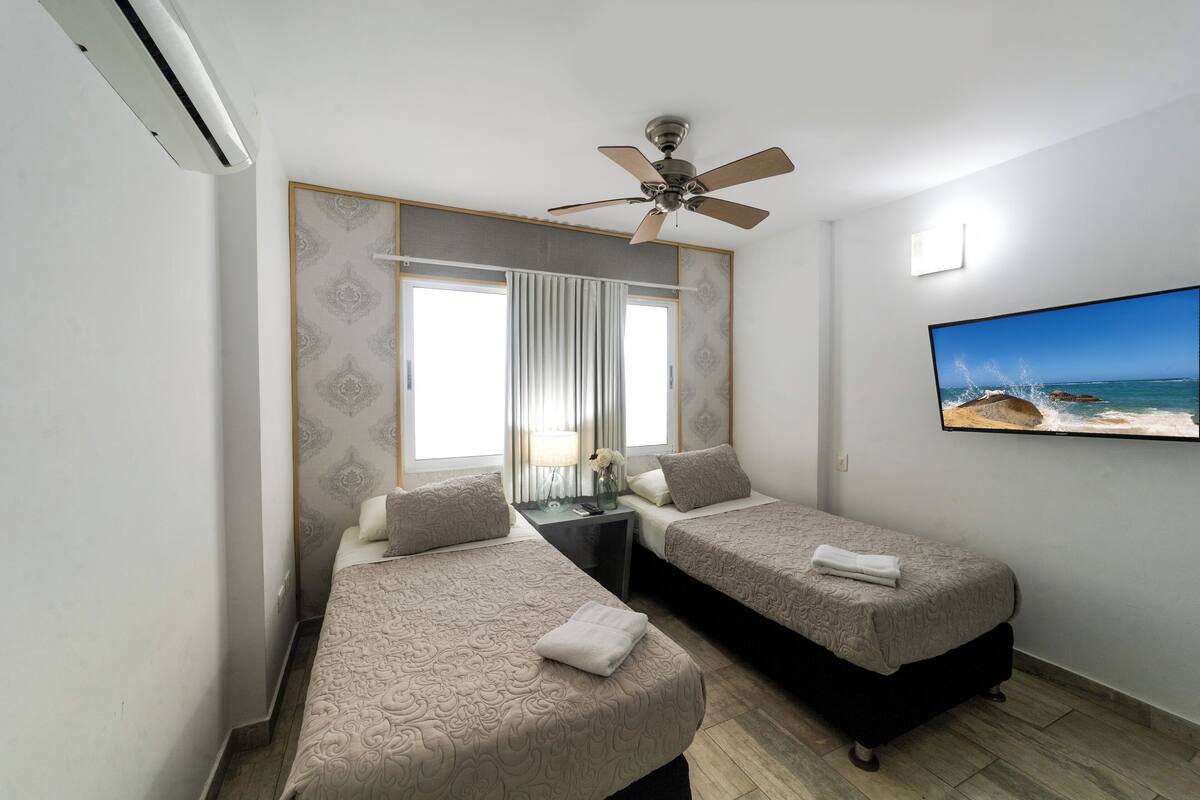 A hotel room with two beds, a ceiling fan, an air conditioner, a window with curtains, a small table with a lamp, and a TV on the wall.