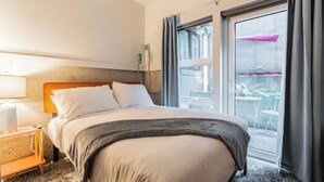 Una habitación de hotel con cama, mesita de noche, lámpara y un amplio ventanal con cortinas.