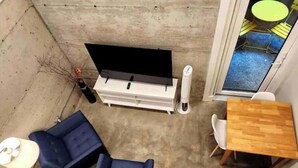 Una stanza con una TV a schermo piatto su un supporto bianco, due poltrone blu, un tavolo in legno e un piccolo tavolino bianco.