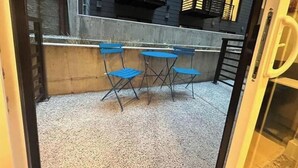 Pieni ulkotila, jossa on pöytä ja neljä tuolia, betoninen reunus sekä taustalla oleva rakennus, jossa on useita ikkunoita ja parvekkeita.