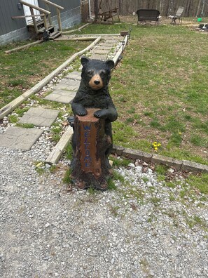 A bear statue with a wooden sign that reads 'WELCOME' in a yard with a gravel path and a wooden fence.