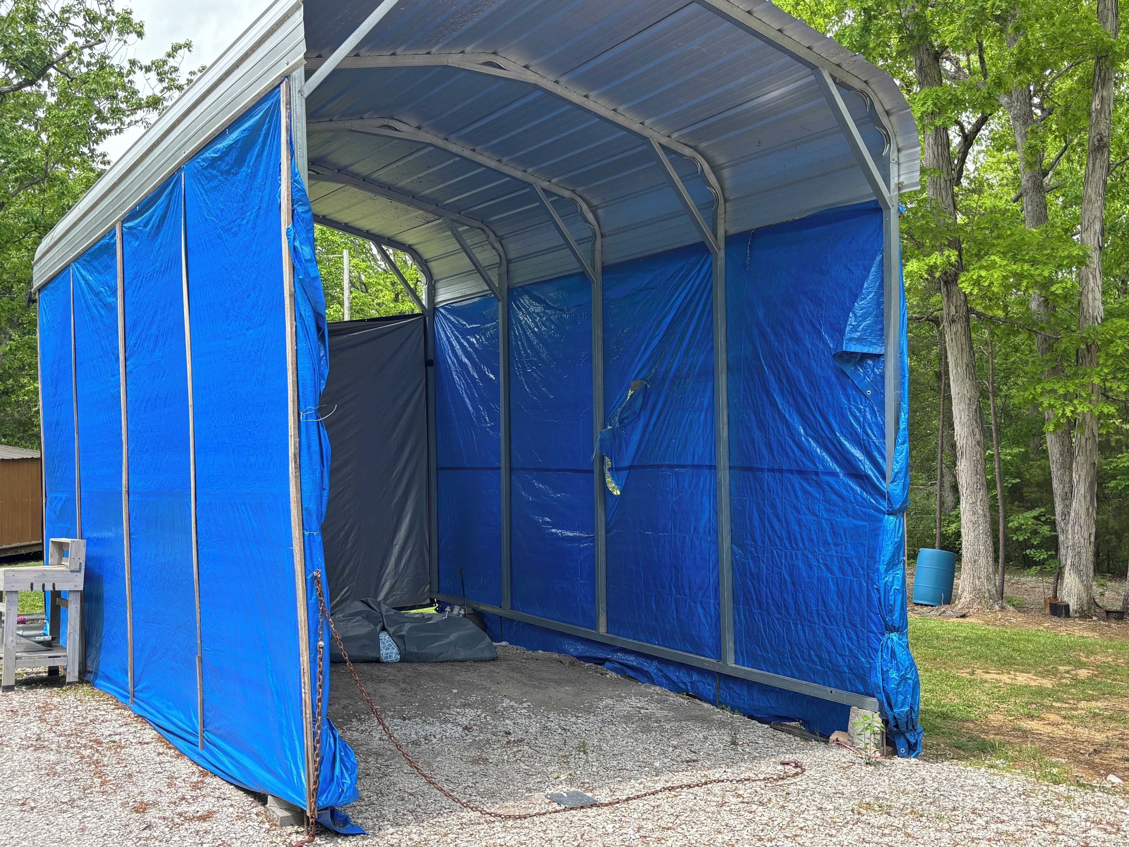 Une structure extérieure abritée, dotée d’un toit en métal et de murs bleus, entourée d’arbres.