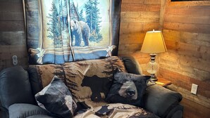 A cozy cabin room with a wooden interior, a window with a forest scene curtain, a lamp, and a quilted bedspread featuring animal prints.