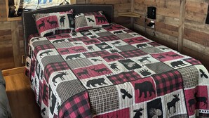 Une chambre confortable avec un lit agrémenté d’une courtepointe à motifs d’élans, un téléviseur fixé au mur diffusant divers services de streaming et un plafond en bois avec un ventilateur de plafond.