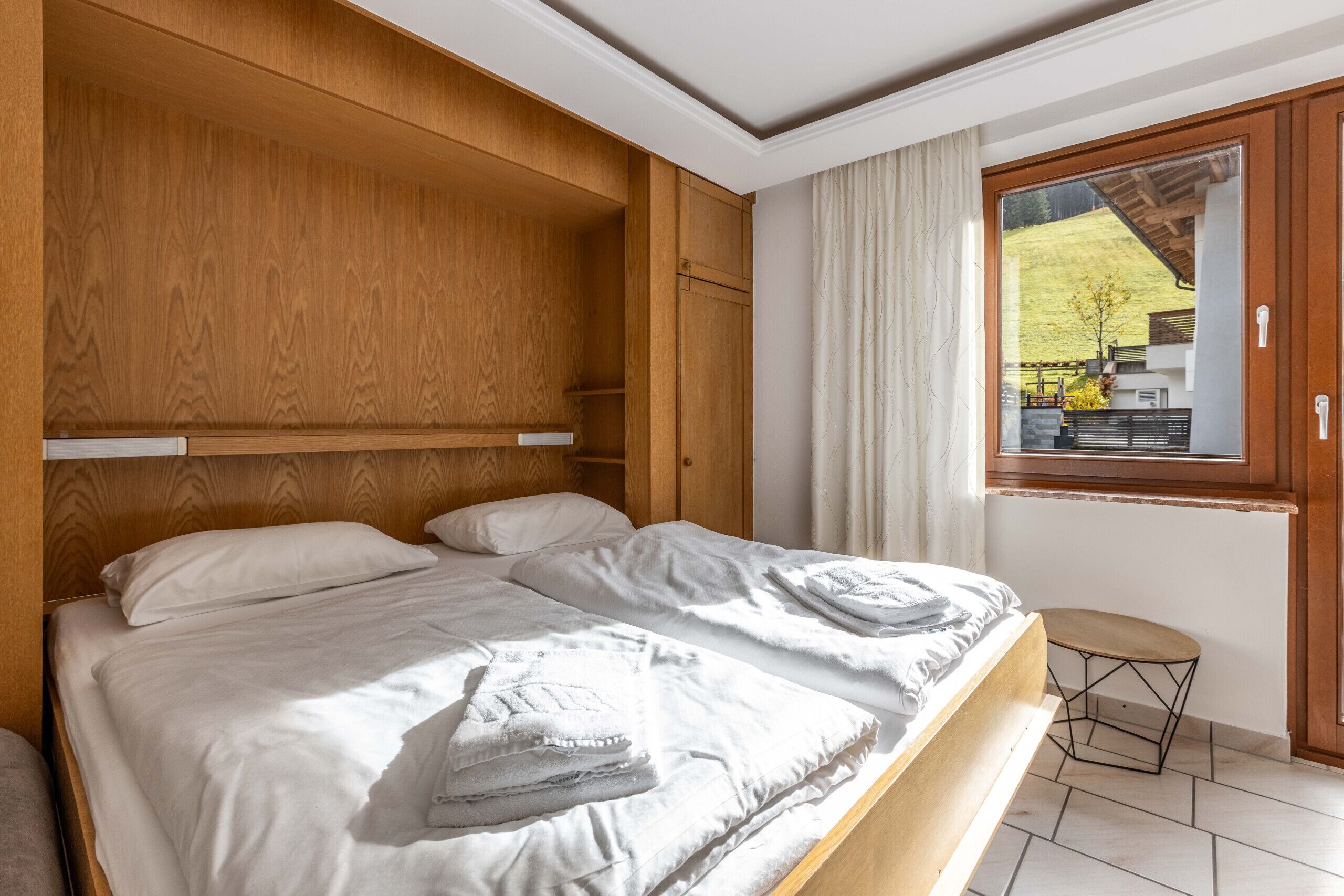 A bedroom with a wooden headboard, two beds with white linens, a small stool, and a window with a view of a green landscape.