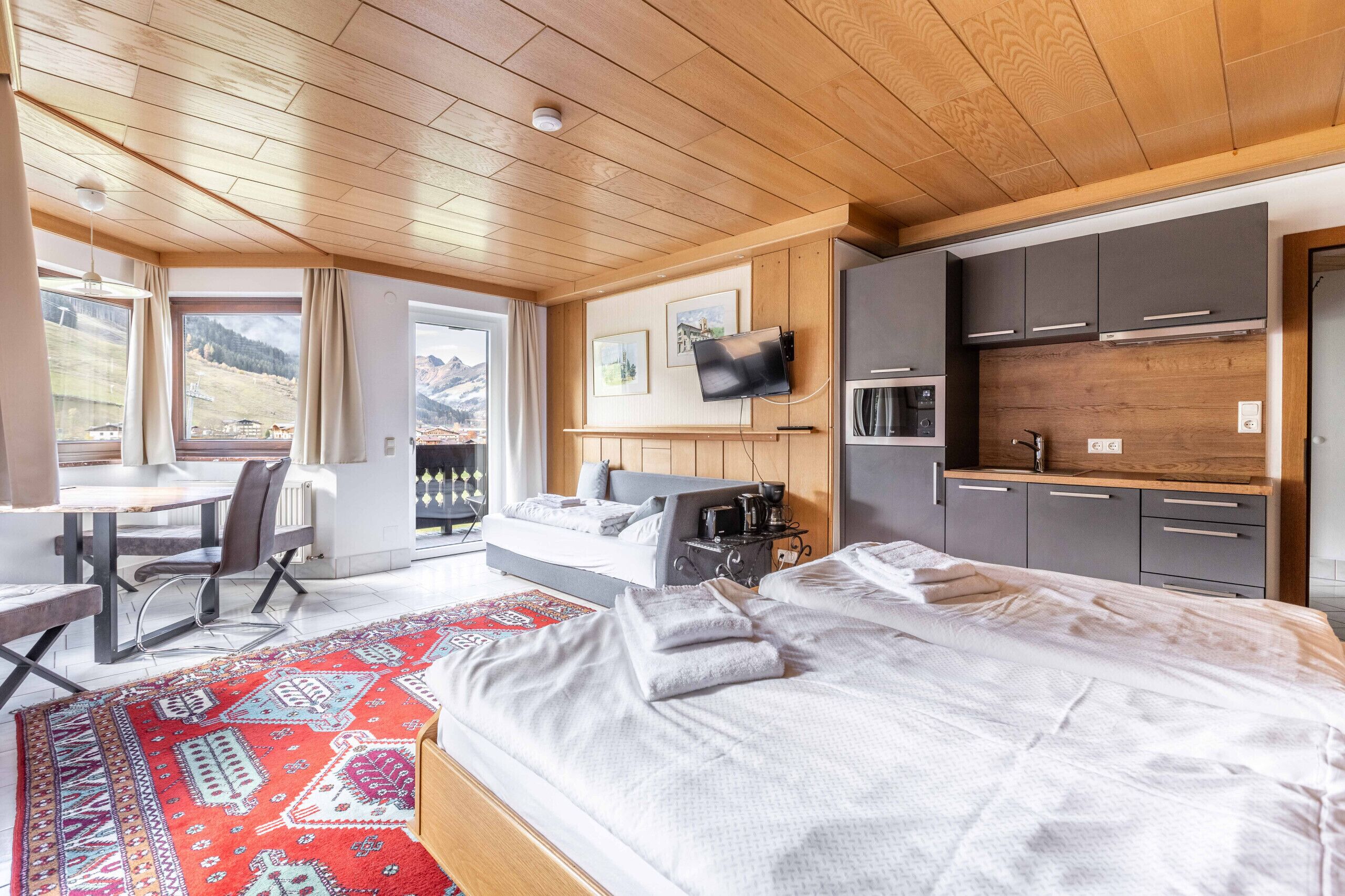 A modern hotel room with a large bed, a desk, a sofa, and a kitchen area.