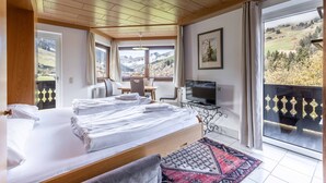 A hotel room with two beds, a TV, a desk, and a balcony with a view of the mountains.