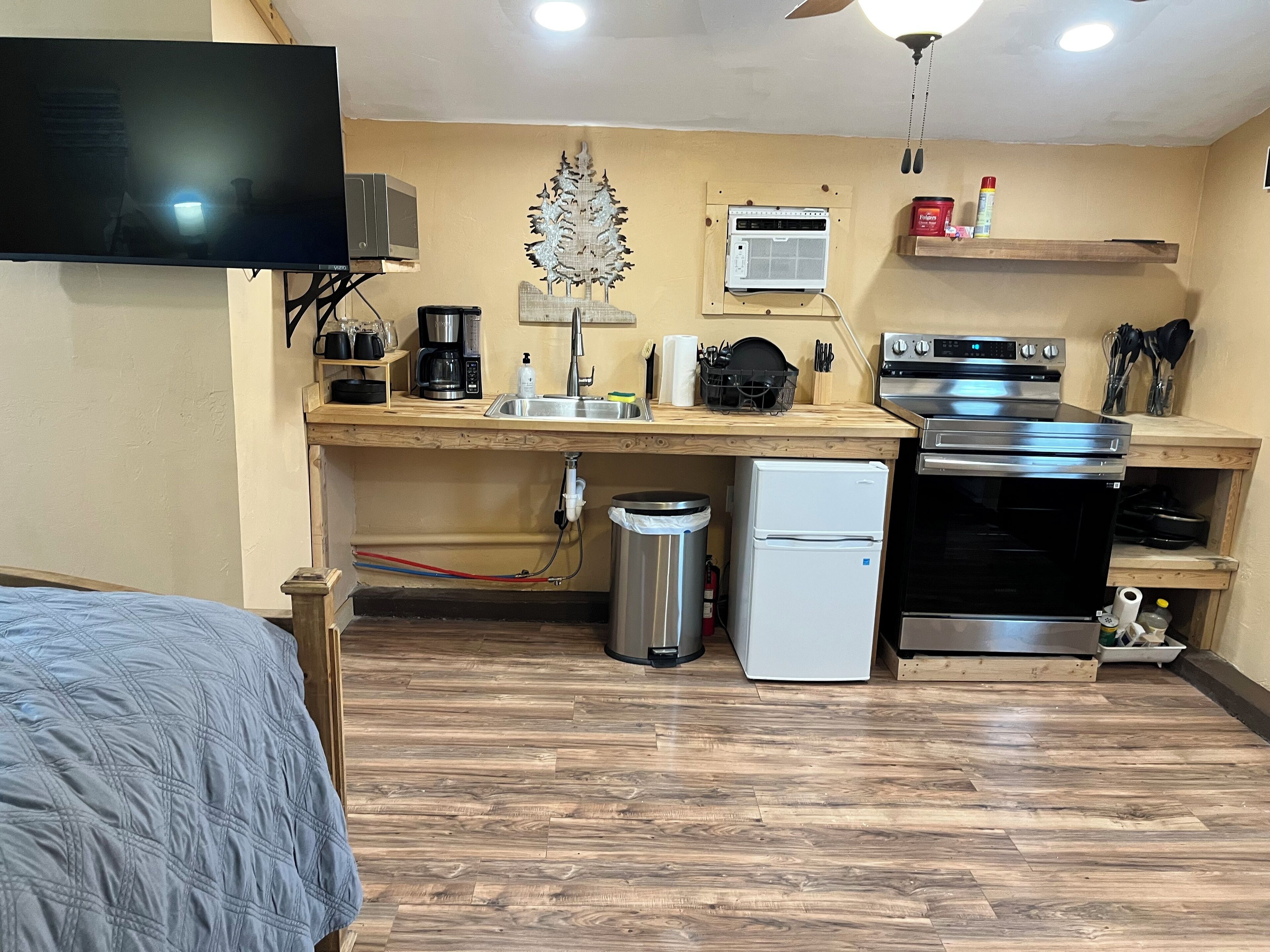 A compact kitchen with a refrigerator, stove, and microwave. There is a wooden countertop with a coffee maker and a sink. A small trash can is visible, and a ceiling fan is mounted above.