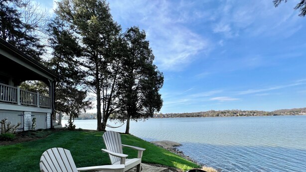 Dos sillas Adirondack sobre un patio de piedra junto a un lago, con un bote de basura metálico cerca.