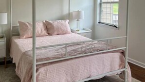 Un lit à baldaquin avec une structure blanche et une literie rose, un ventilateur de plafond, une table de chevet avec une lampe et une fenêtre donnant sur un paysage verdoyant.