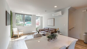 A modern living room with a white sofa, a dining table with chairs, and a large window offering a view of the outside. - Alki Home w Parking, Fast WiFi, Steps to the Beach (Seattle)
