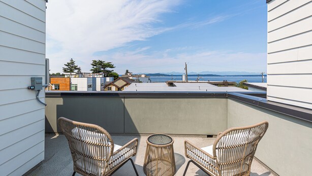 A rooftop terrace with two wicker chairs and a small round table, overlooking a cityscape and a body of water. - Alki Home w Parking, Fast WiFi, Steps to the Beach (Seattle)