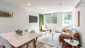 A modern living room with a dining area, large windows, a flat-screen TV, and a green wall accent. - Alki Home w Parking, Fast WiFi, Steps to the Beach (Seattle)
