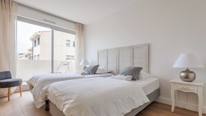 A hotel room with two beds, a chair, a lamp, and a view of buildings outside. - Appartement Limas - Welkeys (Avignon)