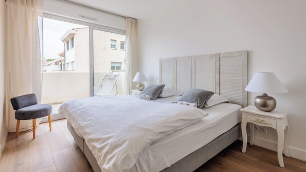A bedroom with a large bed, two bedside tables with lamps, a chair, and a view of buildings through a window. - Appartement Limas - Welkeys (Avignon)