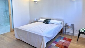 A bedroom with a bed, two bedside tables, a nightstand, a glass shower enclosure, and a colorful rug.