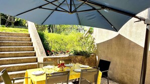 An outdoor dining area with a table set for four, under a large umbrella, surrounded by chairs and a speaker.