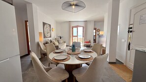 A dining area with a round table, chairs, and a view of the beach through an open door.