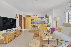 Un salon moderne avec un téléviseur, un canapé et une table basse. Au fond, on aperçoit une cuisine avec des meubles jaunes et un coin repas.