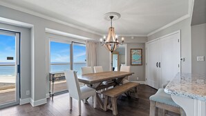 Un comedor con una mesa y sillas de madera, un banco y vistas al océano a través de amplios ventanales.