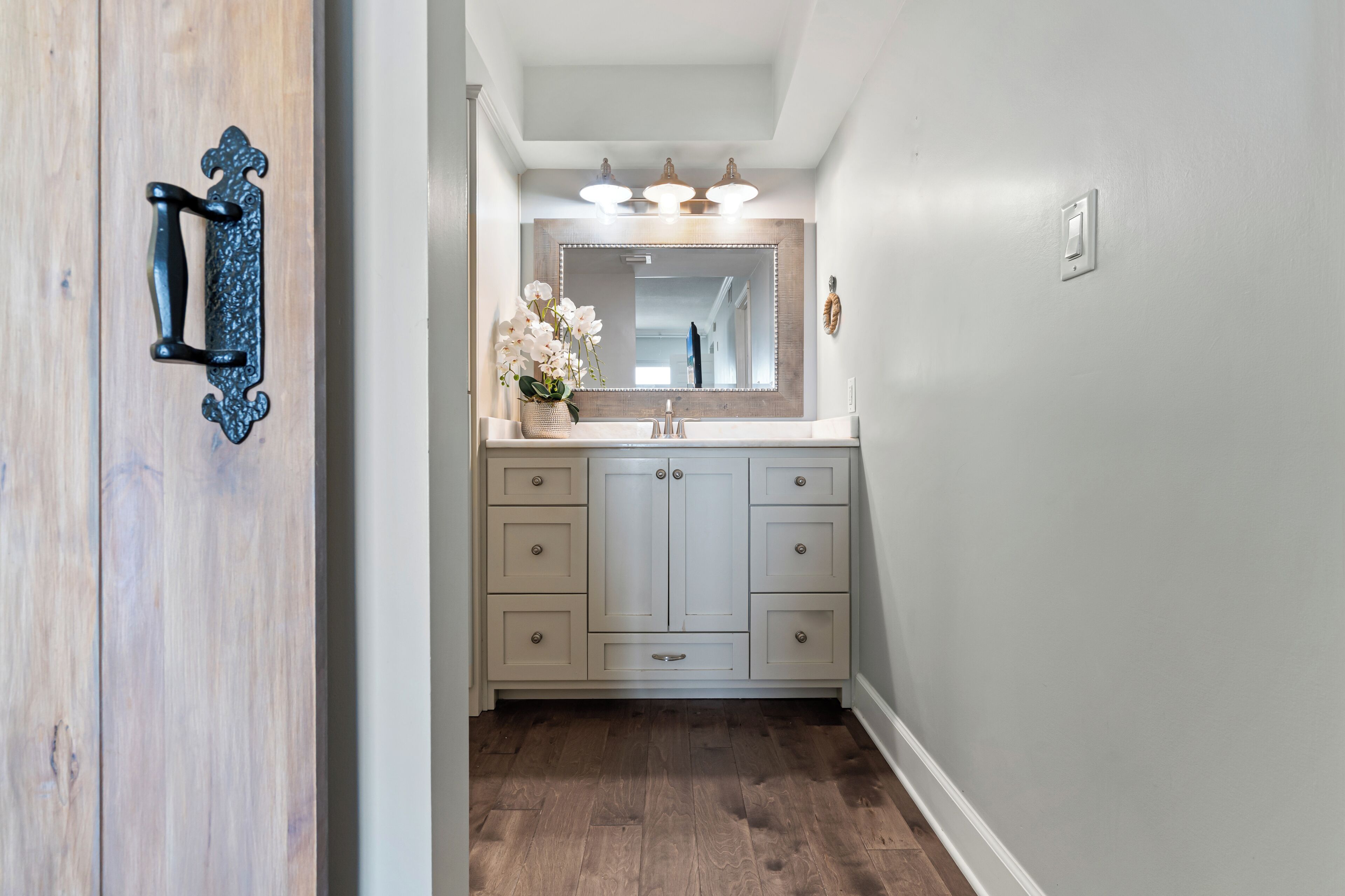 A bathroom with a wooden door, a sink with a mirror, and a light fixture above the mirror.