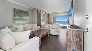 Un salón amplio con un gran ventanal que ofrece vistas a la playa, una mesa de centro de madera y un televisor sobre un mueble de madera.