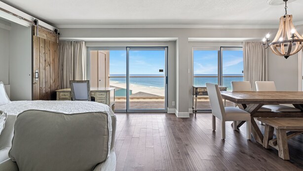 Habitación de hotel con una cama grande, una mesa de comedor de madera y vistas a la playa a través de puertas corredizas de vidrio.