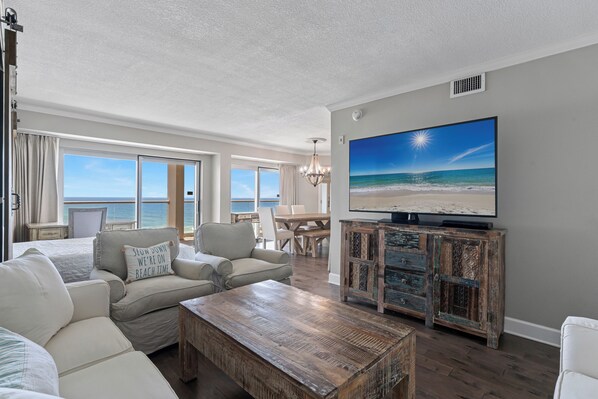 A living room with a large TV, a wooden coffee table, and a view of the ocean.