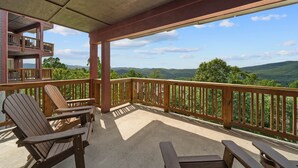 A covered outdoor deck with wooden chairs and a table, overlooking a scenic landscape with mountains and trees. - Mayway Retreat - Echota Club Amenities, Beautiful Mountain Views (Banner Elk)