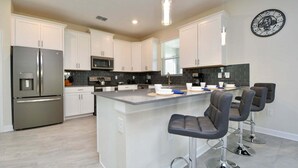 A modern kitchen with white cabinets, stainless steel appliances, a central island with bar stools, and a clock on the wall. - 6BR resort home with private pool, game room & golf access. (Kissimmee)