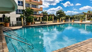 Una piscina con borde de baldosas, rodeada de plantas en macetas y un edificio de varios pisos con balcones.