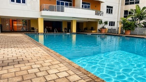 A swimming pool with a tiled deck, a covered seating area, and a building with balconies.