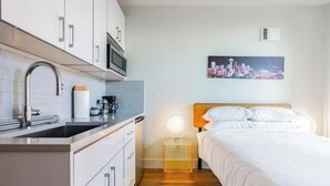 Ein modernes Hotelzimmer mit einer Küchenzeile, einem Bett mit weißer Bettwäsche, einem Nachttisch mit Lampe und einem Stadtbild an der Wand.