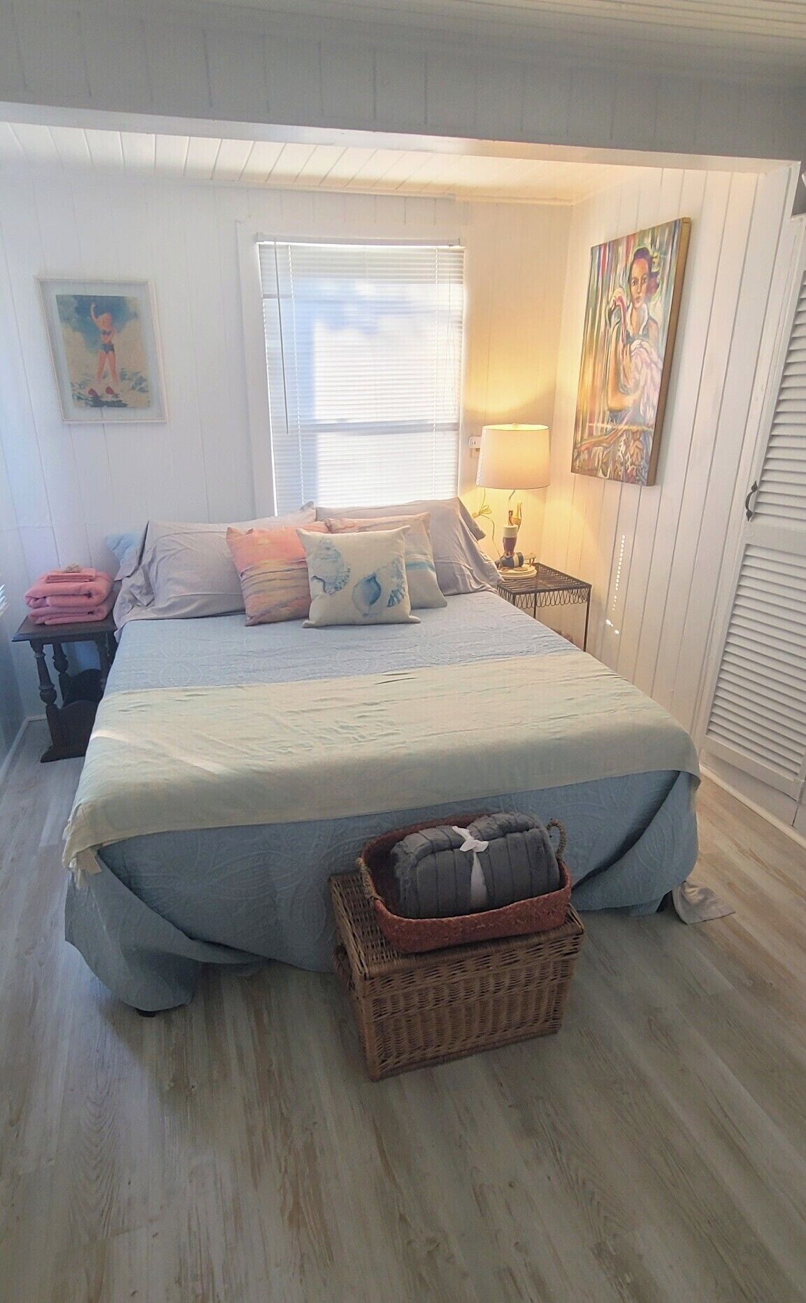 Un dormitorio con cama, mesita de noche, lámpara, cesta de mimbre y ventana con persianas.