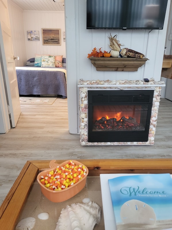 A cozy living room with a fireplace, a television mounted on the wall, and a heart-shaped bowl of candy on the floor.