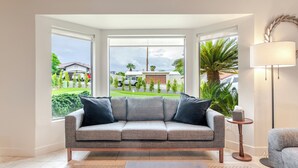 A living room with a grey sofa, a small side table, and a lamp. There are large windows with a view of a house and greenery outside. - Pool, Spa & Hot Tub! Golf Green 5BR near Coachella & Palm Springs-Jamaica Sands (Bermuda Dunes)