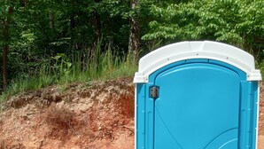 Des toilettes portables avec une porte bleue et des bordures blanches, installées sur une surface de gravier, à proximité d’une barrière métallique.