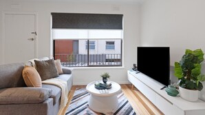 Un salon moderne avec un canapé gris, une table basse ronde, un grand téléviseur à écran plat et une vue sur un immeuble à travers une fenêtre munie d’un store enrouleur foncé.