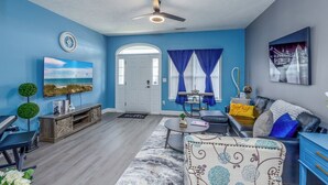 A living room with a blue wall, a ceiling fan, a flat-screen TV, a piano, a sofa, and a coffee table. - Five-Minutes Flyover Cherry Point NC (Havelock)