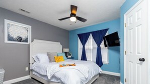 A bedroom with a white bed, a ceiling fan, a TV mounted on the wall, and a window with blue curtains. - Five-Minutes Flyover Cherry Point NC (Havelock)