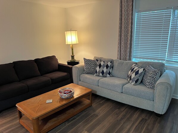 A living room with a dark brown sofa, a light grey sofa with patterned cushions, a wooden coffee table, and a floor lamp.