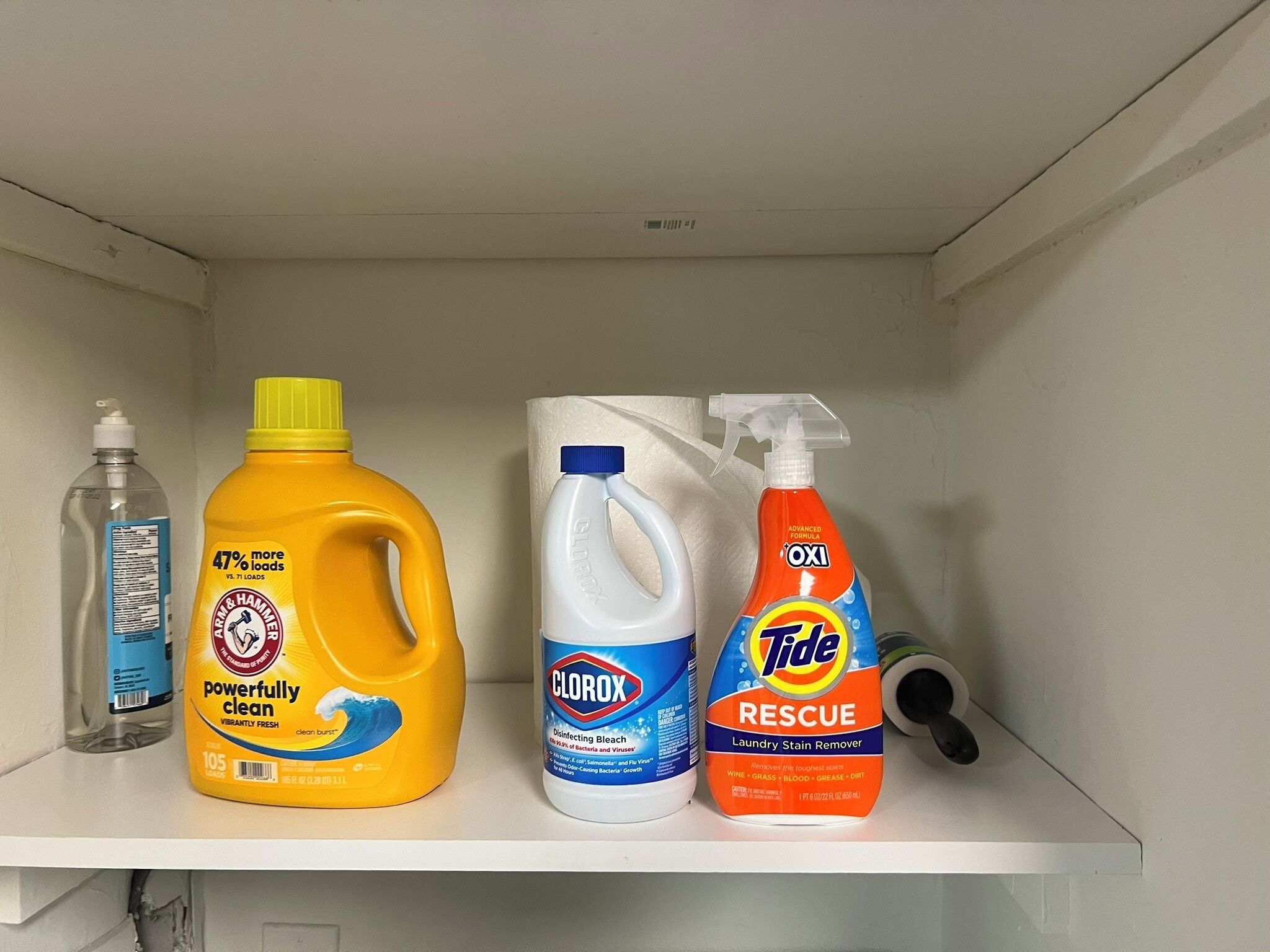 A shelf with three bottles of cleaning products: a yellow bottle of Arm & Hammer Powerfully Clean, a white bottle of Clorox Disinfecting Bleach, and an orange bottle of Tide Rescue Laundry Stain Remover.