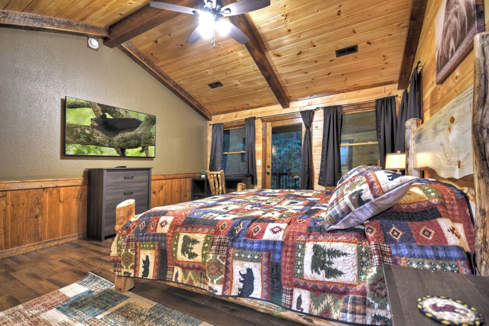 Une chambre confortable, aménagée dans le style d’un chalet, avec un plafond en bois, un grand lit recouvert d’une courtepointe colorée, un bureau avec une chaise et un téléviseur fixé au mur.