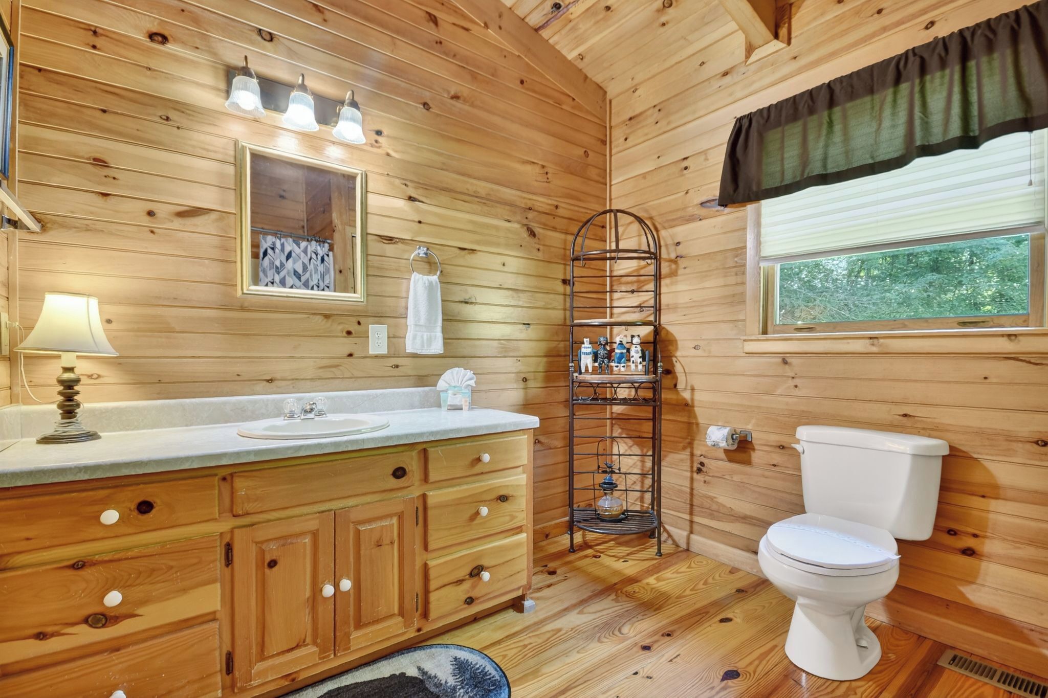 A wooden cabin bathroom with a wooden vanity, a toilet, a shelf with toiletries, and a window with a view of trees.