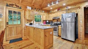 A wooden cabin kitchen with a central island, stainless steel appliances, and a large window overlooking greenery. - Stunning Surroundings, Hot Tub Soaks, S'mores & Nearby Attractions - Our Favorite Place (Blue Ridge)