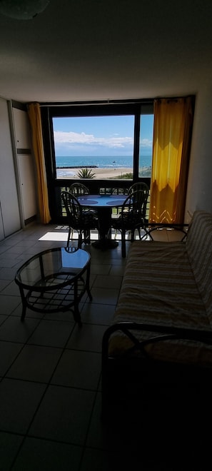 A room with a sofa, a glass table, and a view of the beach through a large window. - Beachfront apartment for 4 people with sea view (Agde)