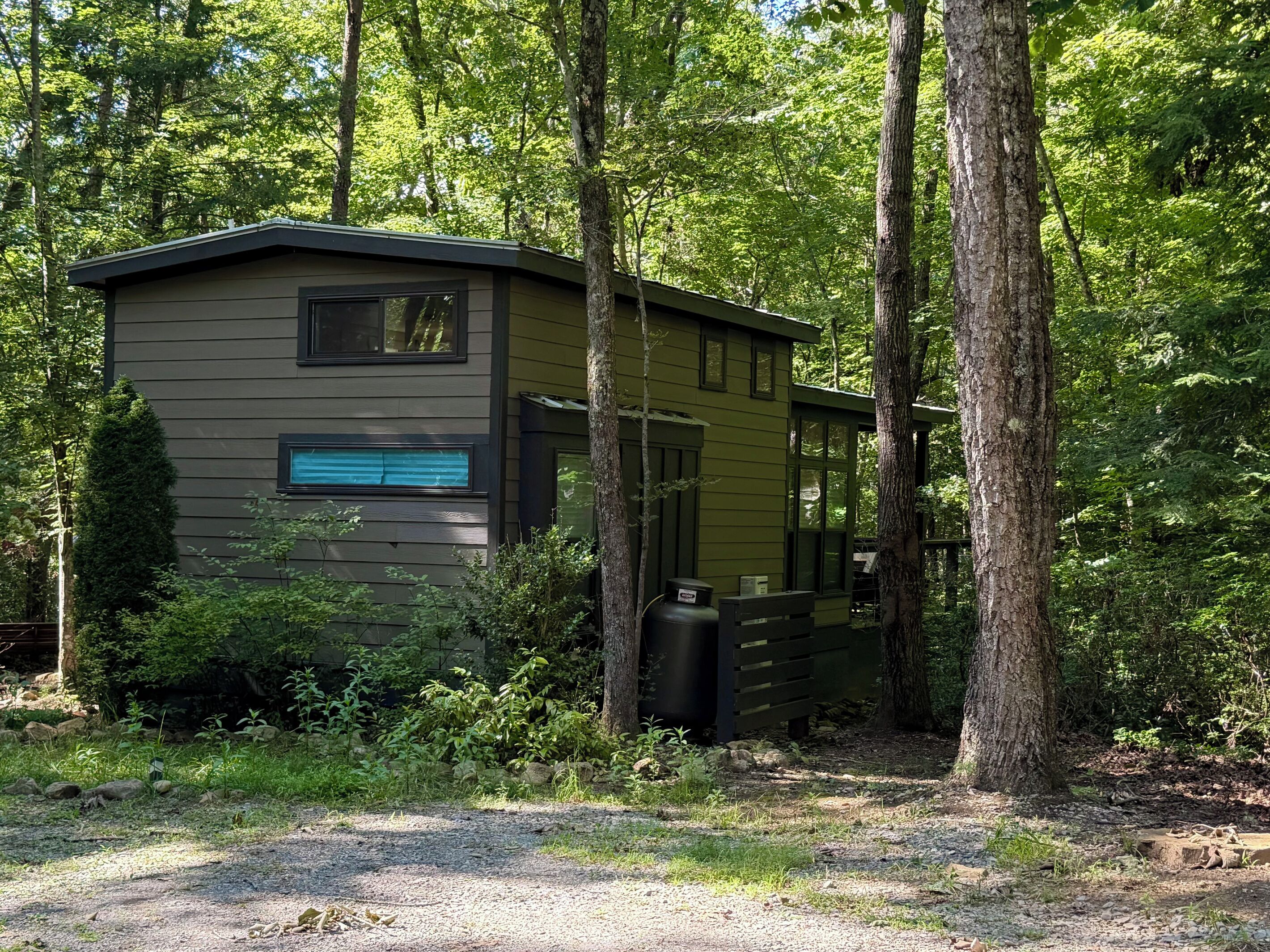 A small, single-story cabin with a porch, surrounded by dense forest.