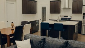 A modern kitchen with dark cabinets, a central island with bar stools, and a dining area with a table and chairs. - Welcome to our Luxury Nest…A home away from home. Your comfort is our culture (Airdrie)