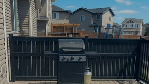 A deck with a grill and a trash can, surrounded by a wooden fence and houses in the background. - Welcome to our Luxury Nest…A home away from home. Your comfort is our culture (Airdrie)