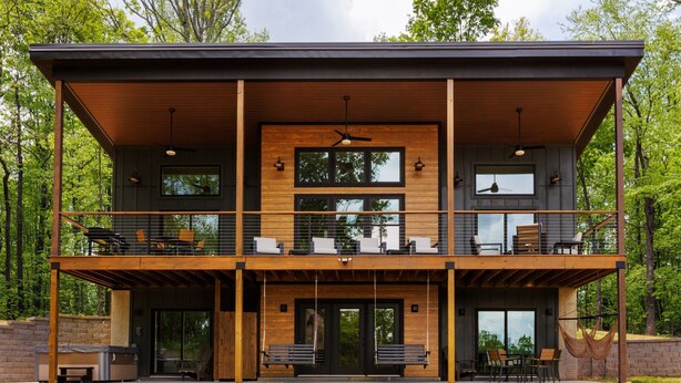 Une maison moderne à deux étages, dotée d’une terrasse en bois, d’un espace salon extérieur et de grandes fenêtres.